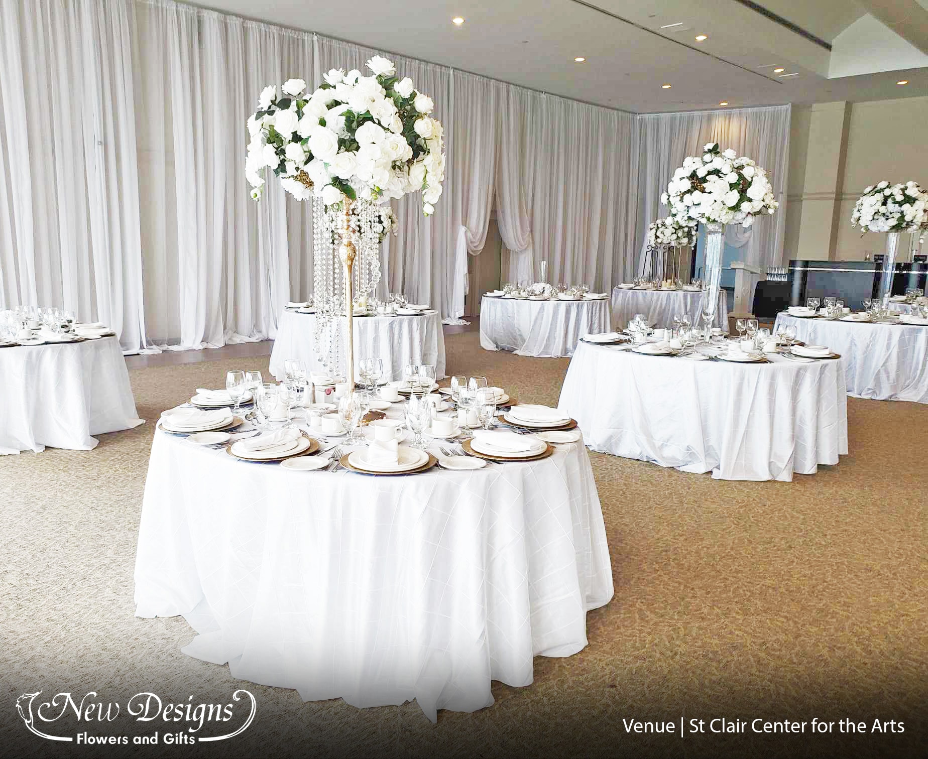 A Reception hall overflowing with Draped white chiffon along the walls and tables. With a Tall Centerpiece Full and compact with White Roses and Orchids, sitting on top A 4 Posted Gold Metal Stand